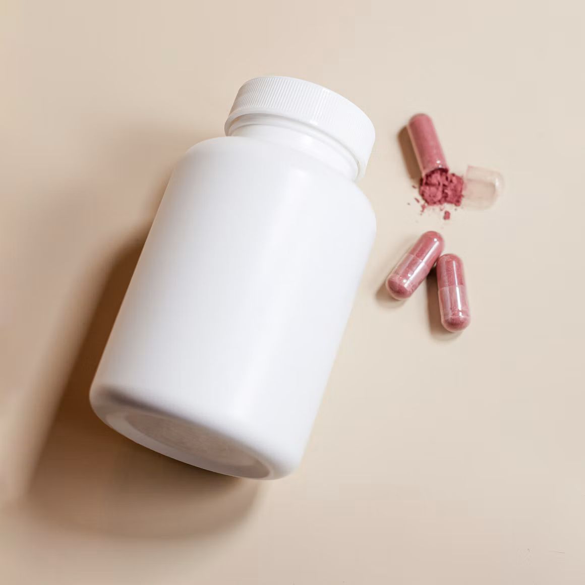 Plain white supplement bottle beside three pink capsules, one of them opened with its powder spilled